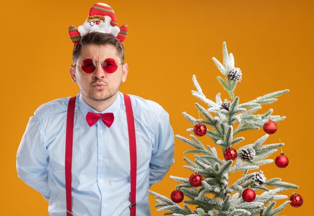 Jonge man met bretels vlinderdas in rand met kerstman en rode bril permanent naast kerstboom camera nauw kijken met ernstig gezicht over oranje achtergrond