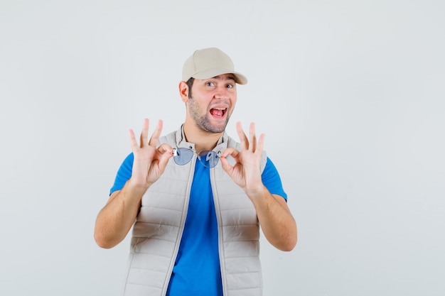 Jonge man met bril in t-shirt, jasje, pet en op zoek gelukkig, vooraanzicht.