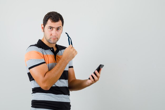 Jonge man met bril terwijl hij mobiele telefoon in t-shirt, bril houdt en voorzichtig kijkt. vooraanzicht. ruimte voor tekst