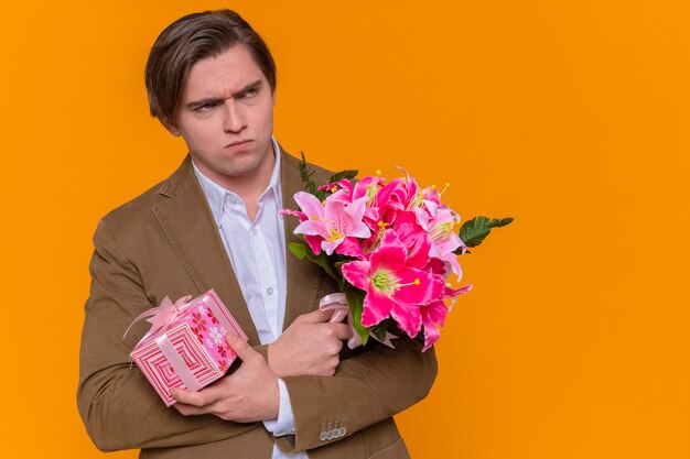 Gratis foto jonge man met cadeau en boeket bloemen opzij kijken met fronsend gezicht gaat feliciteren met internationale vrouwendag die over oranje muur staat