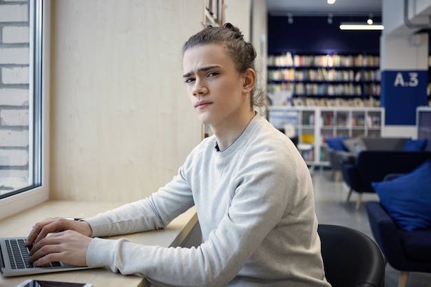 Jonge man met draagbare notebook in café, zittend aan tafel bij het raam en iets te typen, fronsend