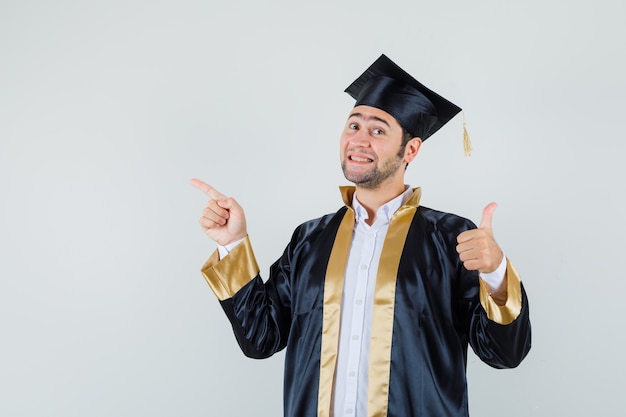 Jonge man met duim omhoog, omhoog in afgestudeerde uniform en op zoek blij, vooraanzicht.