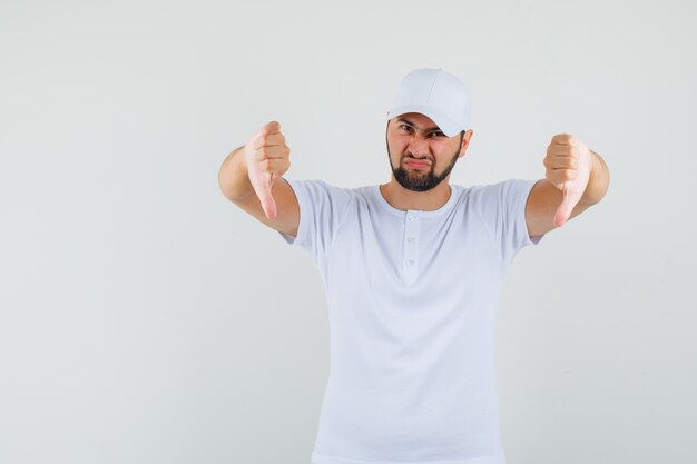 Jonge man met duim omlaag in wit t-shirt, pet en op zoek naar ontevreden, vooraanzicht.