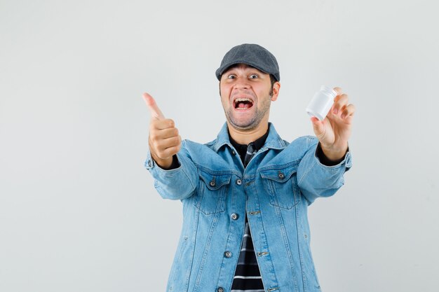 Jonge man met een fles pillen terwijl hij zijn duim in jasje, pet toont en vrolijk kijkt.