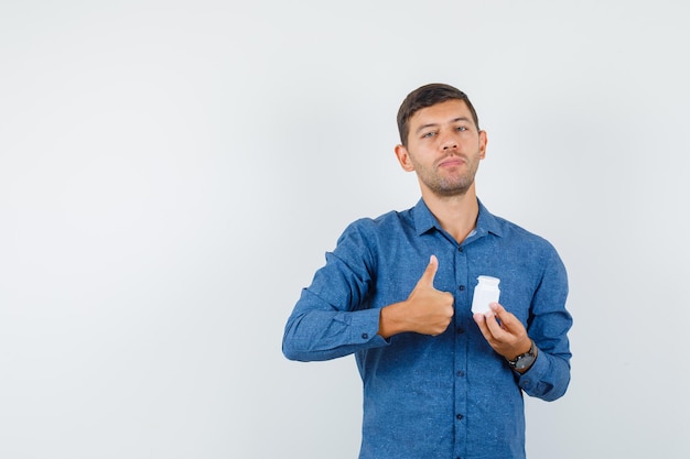 Jonge man met fles pillen met duim omhoog in blauw shirt en tevreden, vooraanzicht.