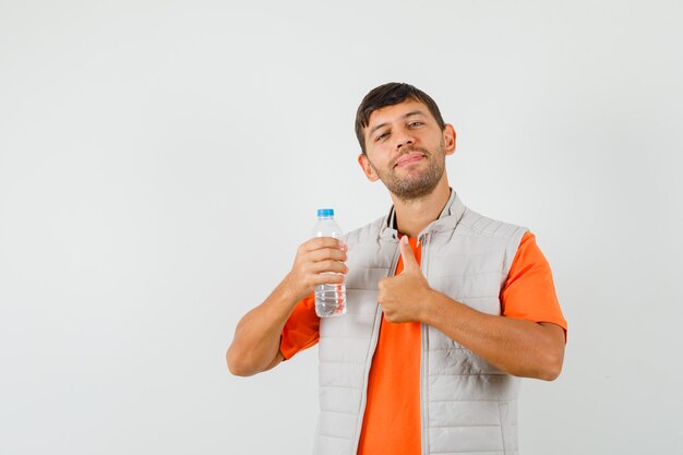 Jonge man met fles water, duim opdagen in t-shirt, jas en op zoek tevreden. vooraanzicht.