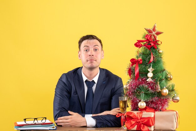 jonge man met gekruiste hand zittend aan de tafel in de buurt van kerstboom en presenteert op geel