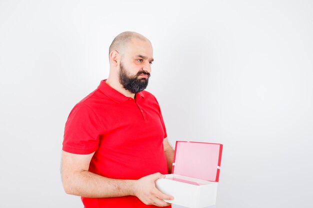 Jonge man met geopende geschenkdoos in rood t-shirt en ontevreden, vooraanzicht.