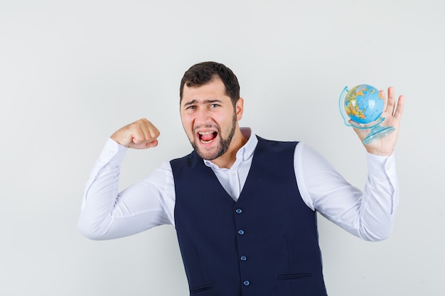 Gratis foto jonge man met globe met winnaar gebaar in shirt, vest en op zoek gelukkig