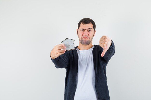 Jonge man met huismodel in de ene hand en duim omlaag in wit t-shirt en zwarte hoodie met rits aan de voorkant en ontevreden kijken. vooraanzicht.