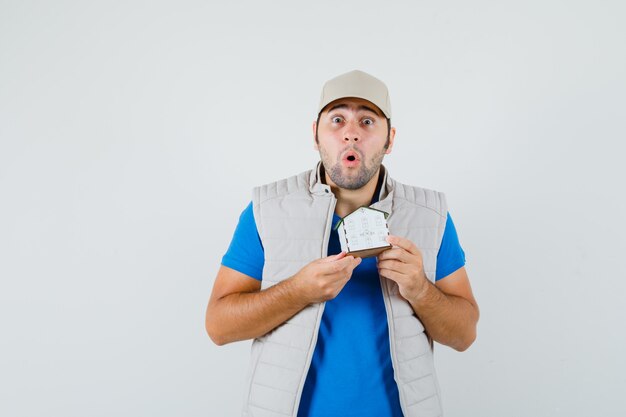 Jonge man met huismodel in t-shirt, jasje, pet en op zoek gealarmeerd. vooraanzicht.