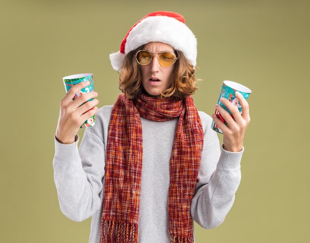 Jonge man met kerst kerstmuts en gele bril met warme sjaal om zijn nek met kleurrijke papieren bekers op zoek verward met twijfels staande over groene achtergrond