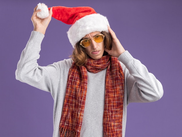 Jonge man met kerst-kerstmuts en gele bril met warme sjaal om zijn nek op zoek vrolijk en blij aanraken van zijn hoed tong uitsteekt staande over paarse achtergrond