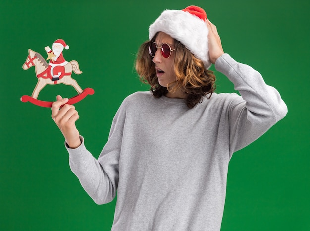Gratis foto jonge man met kerst kerstmuts en rode bril met kerst speelgoed kijken verward met de hand op zijn hoofd staande over groene achtergrond
