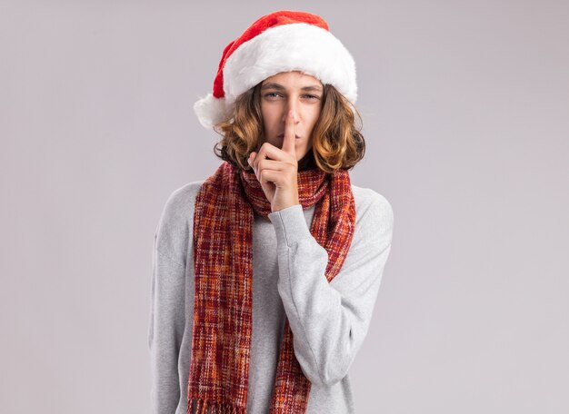 Jonge man met kerst kerstmuts met warme sjaal om zijn nek op zoek stilte gebaar met vinger op lippen kijken