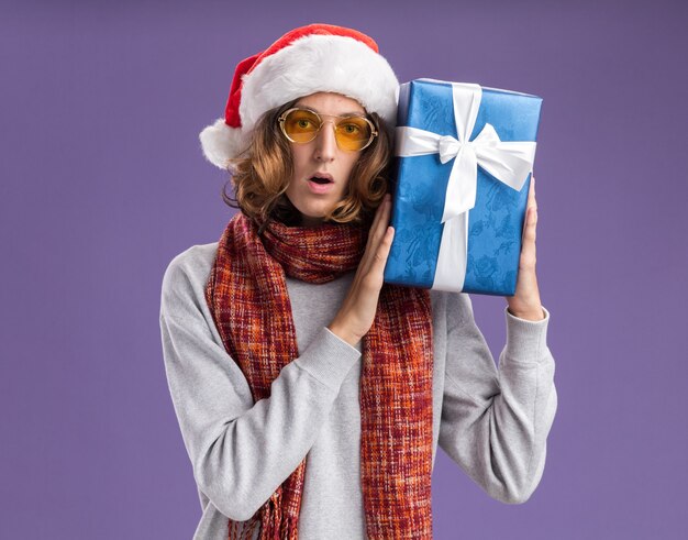 Jonge man met kerstmuts en gele bril met warme sjaal om zijn nek met kerstcadeau verrast over paarse muur