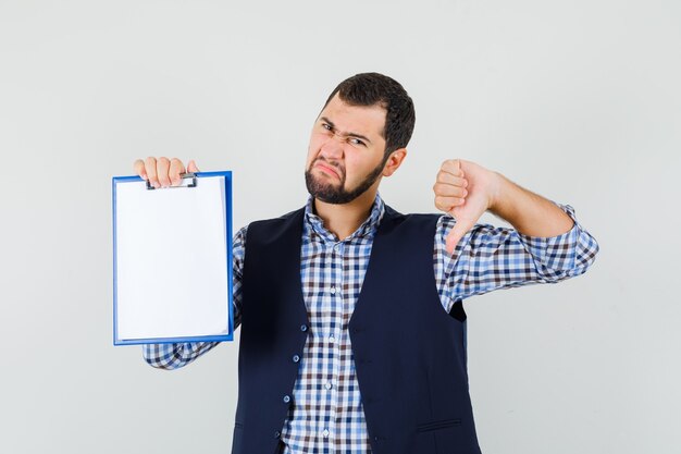 Jonge man met klembord met duim omlaag in shirt, vest en op zoek naar ontevredenheid, vooraanzicht.