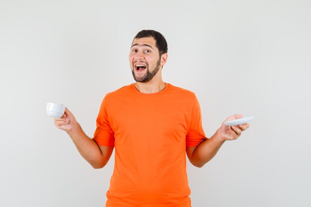 Jonge man met kop en schotel in oranje t-shirt en vrolijk, vooraanzicht.