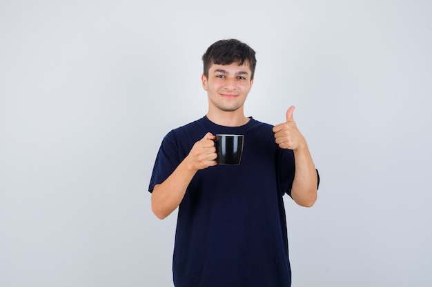 Jonge man met kopje thee, duim opdagen in zwart t-shirt en op zoek vrolijk. vooraanzicht.