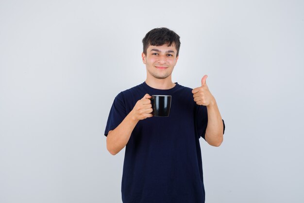 Jonge man met kopje thee, duim opdagen in zwart t-shirt en op zoek vrolijk. vooraanzicht.