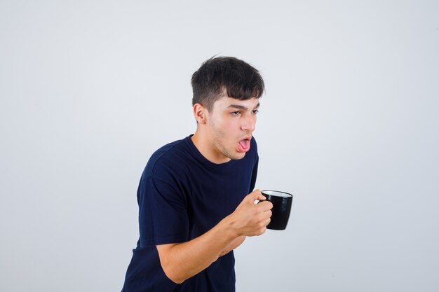 Jonge man met kopje thee terwijl misselijk gevoel in zwart t-shirt en onwel kijken. vooraanzicht.