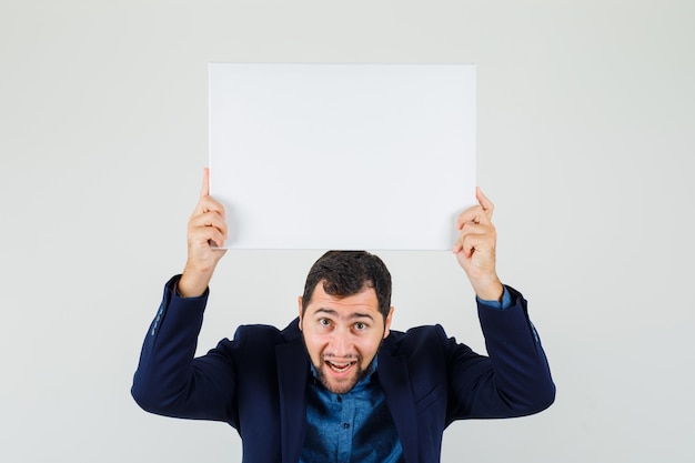 Jonge man met leeg canvas boven het hoofd in shirt, jasje en op zoek gelukkig, vooraanzicht.