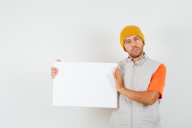 Jonge man met leeg canvas in t-shirt, jasje, hoed en op zoek kalm, vooraanzicht.