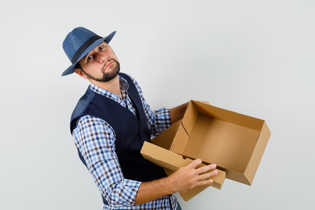 Jonge man met lege kartonnen doos in shirt, vest, hoed en op zoek verdrietig.