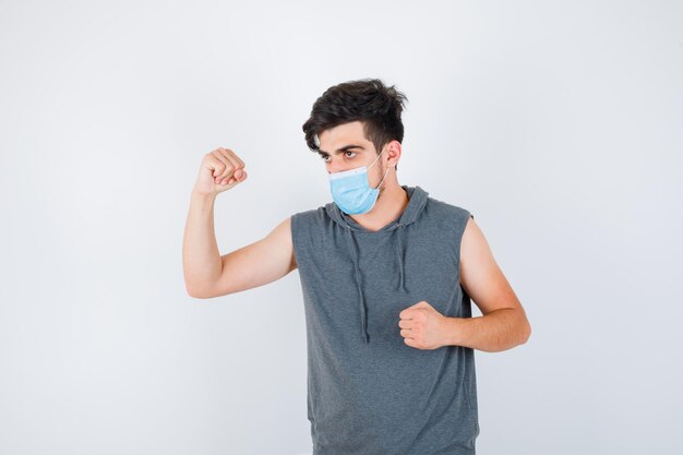 Jonge man met masker terwijl hij spieren toont en vuist in grijs t-shirt balt en er serieus uitziet?