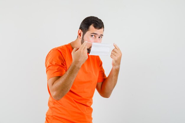 Jonge man met medisch masker in oranje t-shirt en op zoek voorzichtig, vooraanzicht.