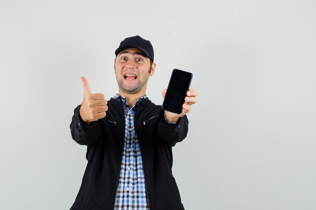 Jonge man met mobiele telefoon, duim opdagen in shirt, jasje, pet en vrolijk kijken.