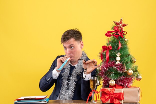 Jonge man met noisemaker zittend aan tafel in de buurt van kerstboom en presenteert op geel