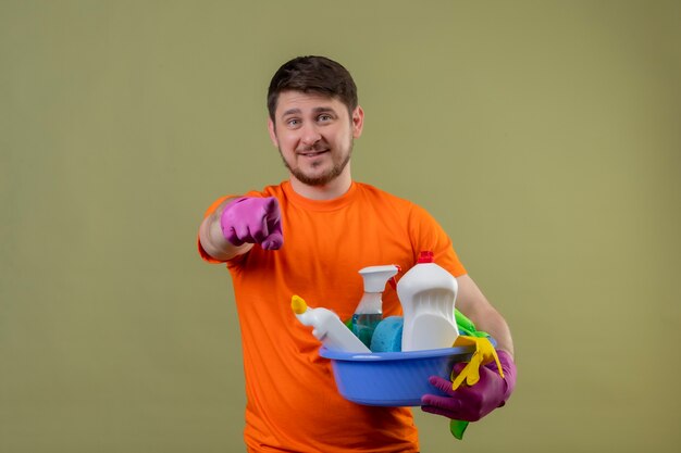 Jonge man met oranje t-shirt en rubberen handschoenen met bekken met schoonmaakgereedschap glimlachend positief en blij wijzend met vinger naar camera staande over groene muur 2