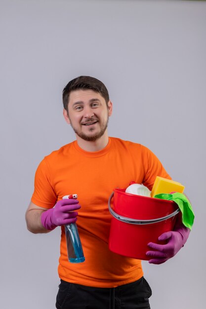 Jonge man met oranje t-shirt en rubberen handschoenen met emmer met reinigingsgereedschap en reinigingsspray