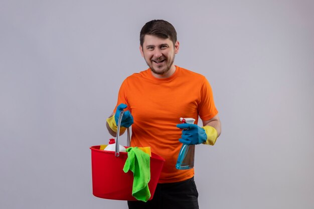 Jonge man met oranje t-shirt en rubberen handschoenen met emmer met reinigingsgereedschap en reinigingsspray