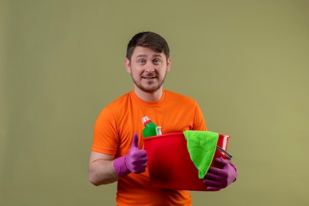 Jonge man met oranje t-shirt en rubberen handschoenen met emmer met reinigingsgereedschap