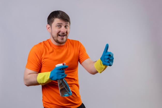 Jonge man met oranje t-shirt en rubberen handschoenen met schoonmaak spray glimlachend vrolijk positief