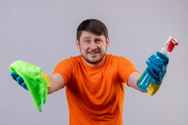 Jonge man met oranje t-shirt en rubberen handschoenen met schoonmaakspray en deken glimlachend vrolijk positief