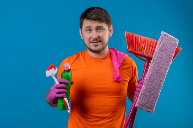 Jonge man met oranje t-shirt en rubberen handschoenen met zwabber en schoonmaakmiddelen met sceptische uitdrukking