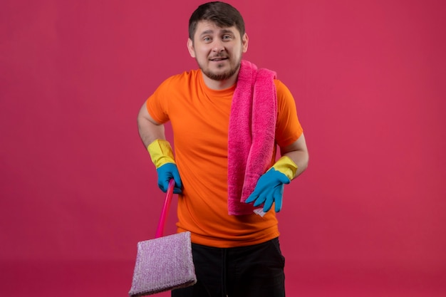 Jonge man met oranje t-shirt en rubberen handschoenen met zwabber met opgeheven arm als het stellen van een vraag staande over roze muur