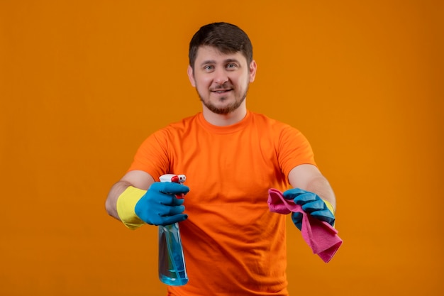 Jonge man met oranje t-shirt met schoonmaakspray en tapijt glimlachend vrolijk positief en gelukkig kijken naar camera klaar om schoon te maken staande over oranje achtergrond