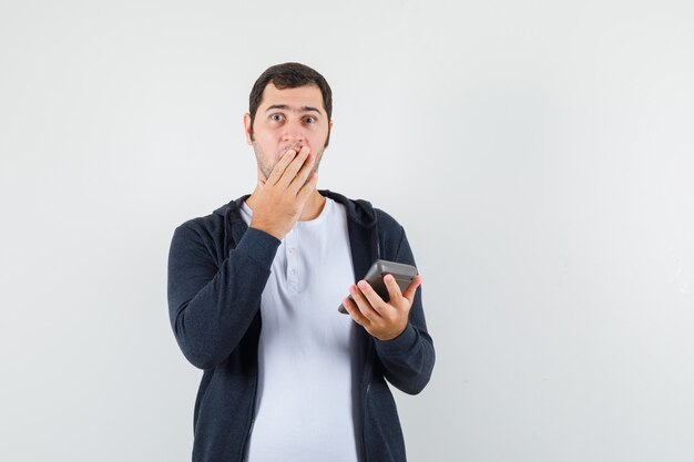 Jonge man met rekenmachine, mond bedekt met hand in wit t-shirt en zwarte hoodie met ritssluiting aan de voorkant en op zoek verrast, vooraanzicht.