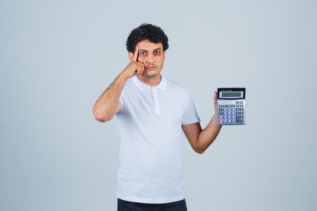 Jonge man met rekenmachine terwijl hij in een wit t-shirt denkt en er verstandig uitziet, vooraanzicht.