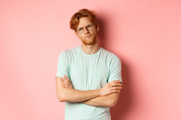 Jonge man met rood haar en baard, met bril en t-shirt, armen gekruist op de borst, fronsend terwijl hij met sceptische en twijfelachtige uitdrukking staart, staande over roze achtergrond.