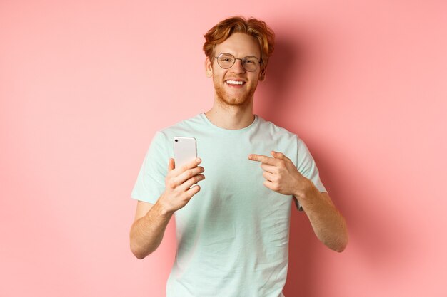 Jonge man met rood haar en baard, met t-shirt en bril, glimlachend terwijl hij met de vinger naar de smartphone wijst, raadt online promotie aan, roze achtergrond.