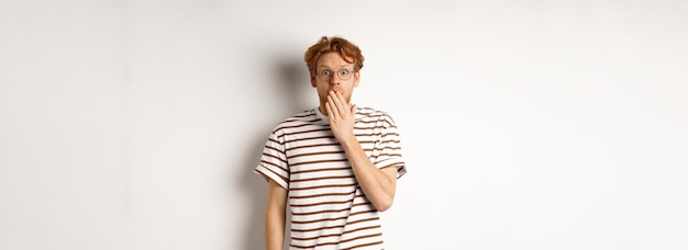 Gratis foto jonge man met rood haar en bril hoort schokkend nieuws of roddelt over mond en staart naar camera