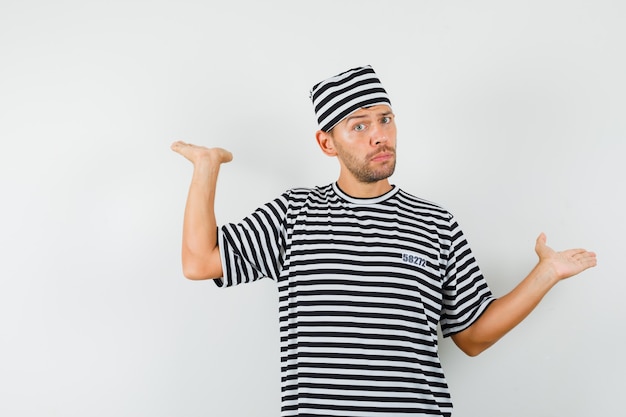 Gratis foto jonge man met schalen gebaar in gestreept t-shirt, hoed en besluiteloos op zoek.
