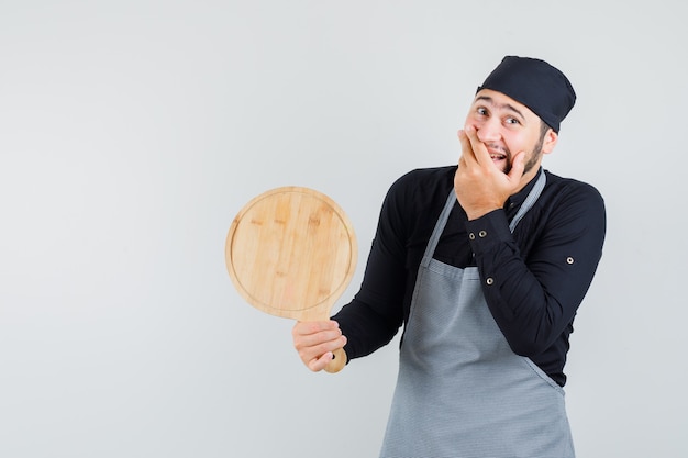 Jonge man met snijplank met hand op mond in shirt, schort en op zoek gelukkig. vooraanzicht.