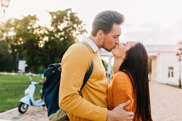 Jonge man met stijlvol kapsel zoenen mooie brunette meisje met gesloten ogen. Portret van een geïnspireerde mooie dame die geniet van een date in de buitenlucht met een vriend in oranje kleding.