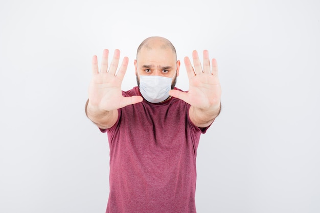 Jonge man met stopgebaar in roze t-shirt, masker en serieus, vooraanzicht.